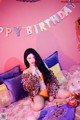 A woman sitting on a bed with balloons and a birthday cake.